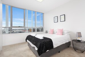 Une chambre moderne avec une grande fenêtre, un lit recouvert de draps blancs, un plaid noir et une table de chevet avec une lampe.
