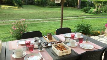Une table de jardin, dressée avec des assiettes, des tasses et un panier de viennoiseries, entourée de chaises.