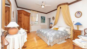 Une chambre à coucher comprenant un lit, une table de chevet, une table avec une lampe, un ventilateur de plafond et une grande fenêtre avec des rideaux.