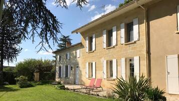 Un bâtiment en pierre à deux étages, doté de volets blancs, une terrasse pavée avec des chaises rouges et une pelouse bien entretenue.
