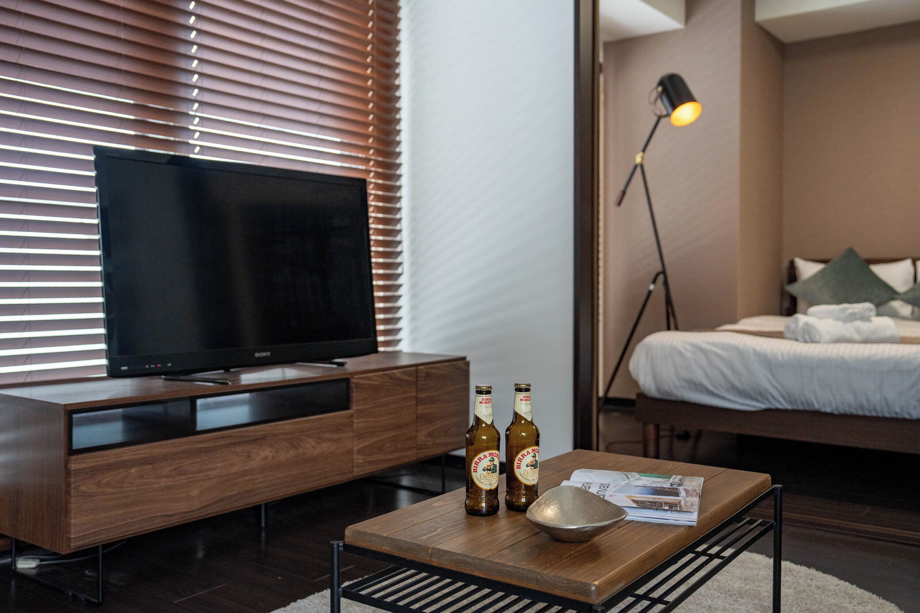 Een moderne hotelkamer met een flatscreen televisie op een houten tv-meubel, een nachtkastje met twee bierflesjes en een schaal, een bed met witte beddengoed, en een vloerlamp.