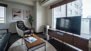 Ett modernt vardagsrum med en soffa, en massagefåtölj, ett soffbord med tidningar, en tv på ett träställ och ett stort fönster med utsikt över byggnader.