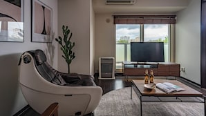 Ett modernt vardagsrum med en fåtölj som kan lutas bakåt, en platt-TV, ett soffbord med tidningar och flaskor, och ett stort fönster med utsikt över staden.