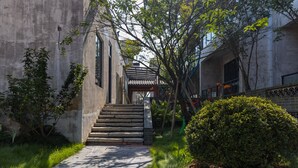 A narrow pathway leading to a traditional wooden structure with a tiled roof, surrounded by greenery and a concrete building. - Wuyuan Wunvzhou Wuxiandao Luxury Hotel  (Shangrao)