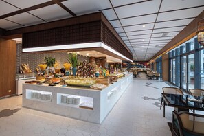 A spacious hotel lobby with a central buffet area, large windows, and a modern ceiling design.