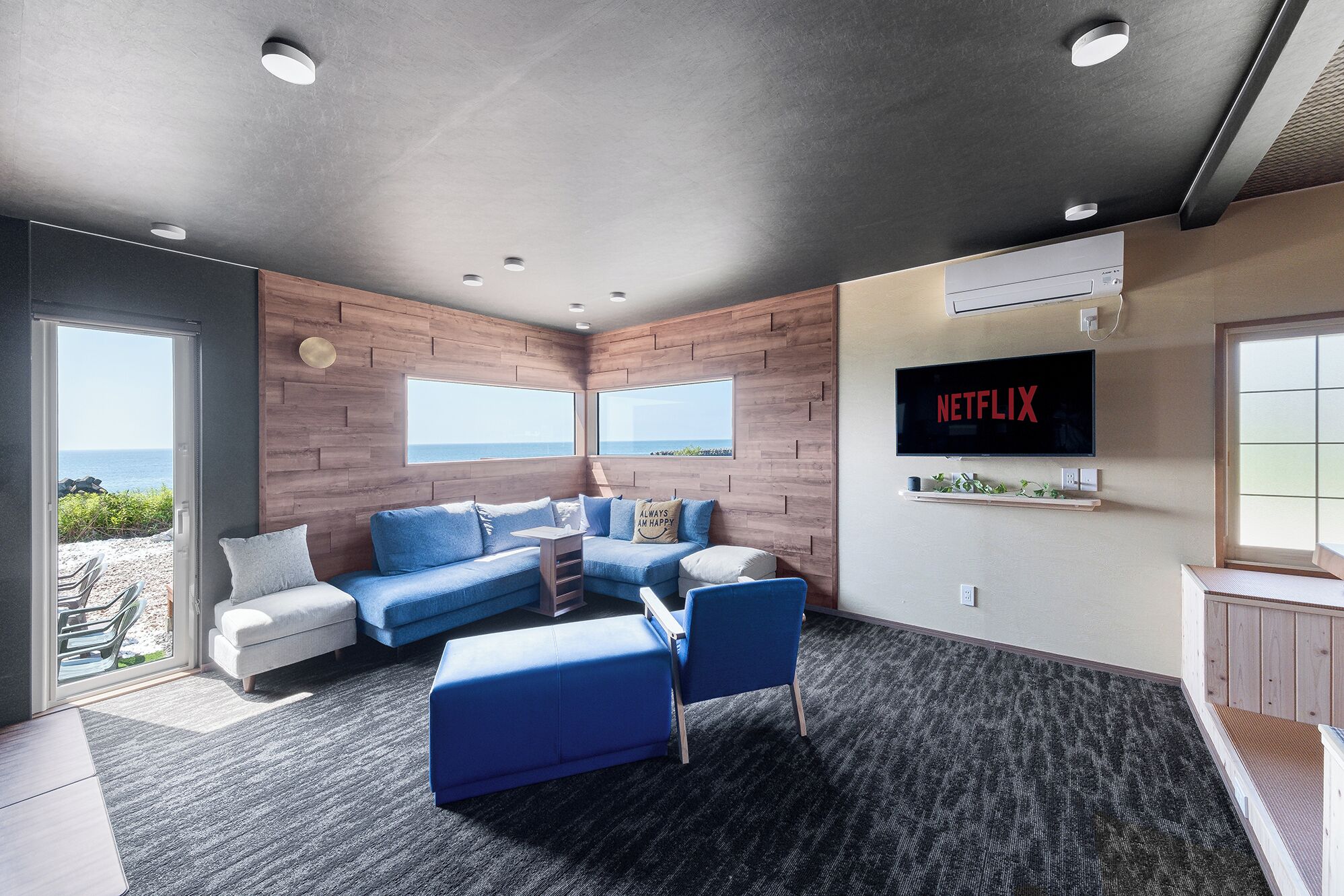 A modern living room with a large TV, comfortable seating, and a view of the sea.