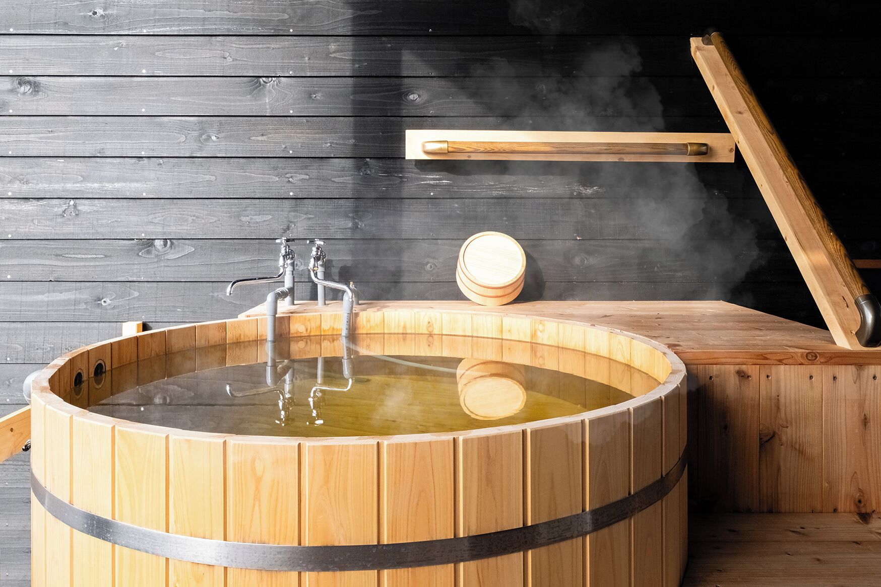 A wooden hot tub with a metal faucet and a wooden bench with a cup.
