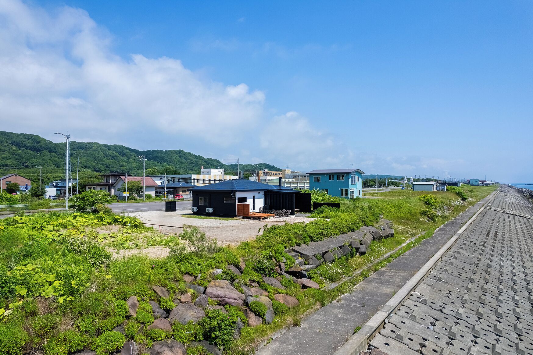 A coastal area with a paved walkway, a small building with a blue roof, and a hillside in the background.