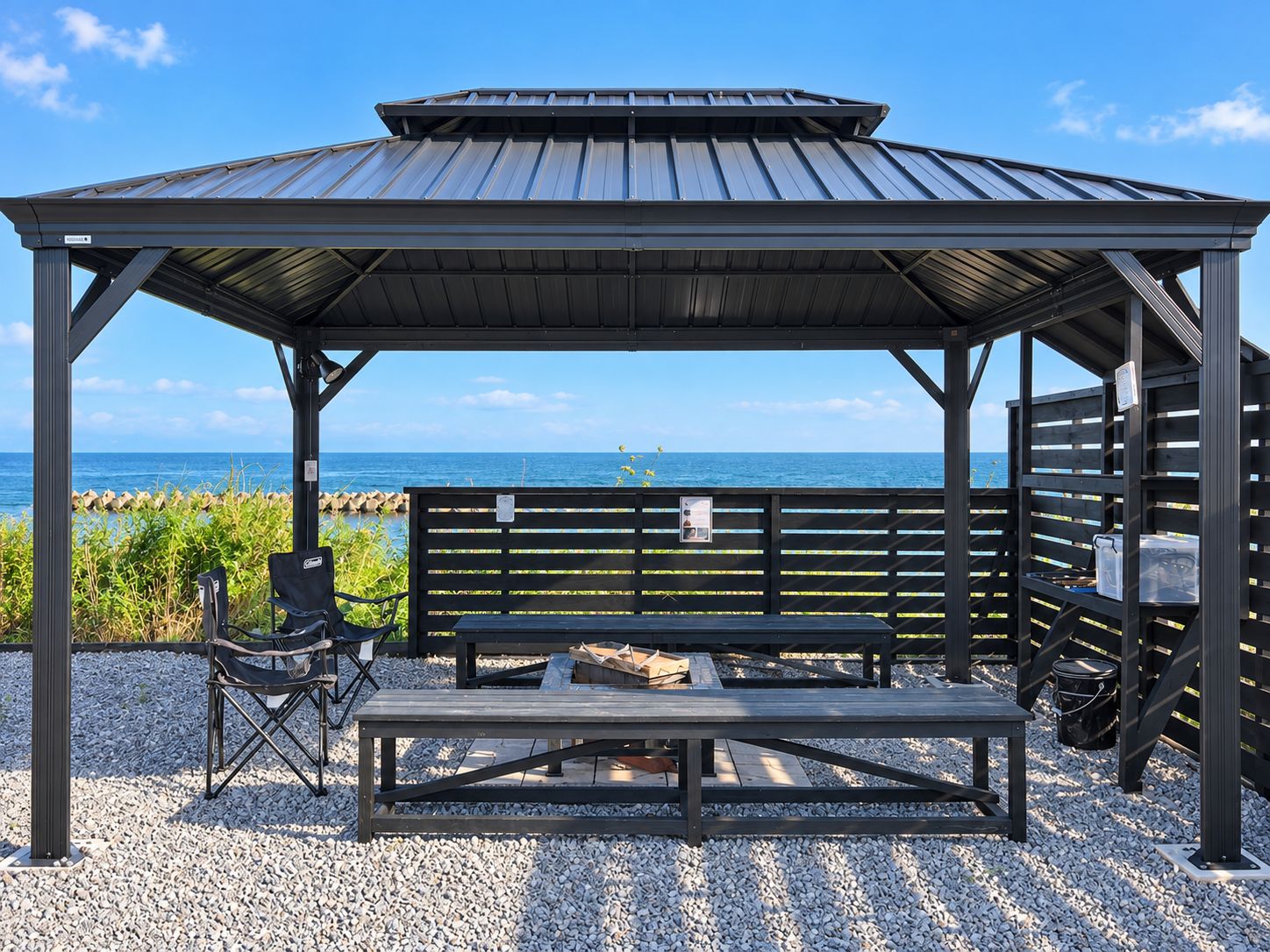 A covered outdoor seating area with a bench and a grill, overlooking a beach.