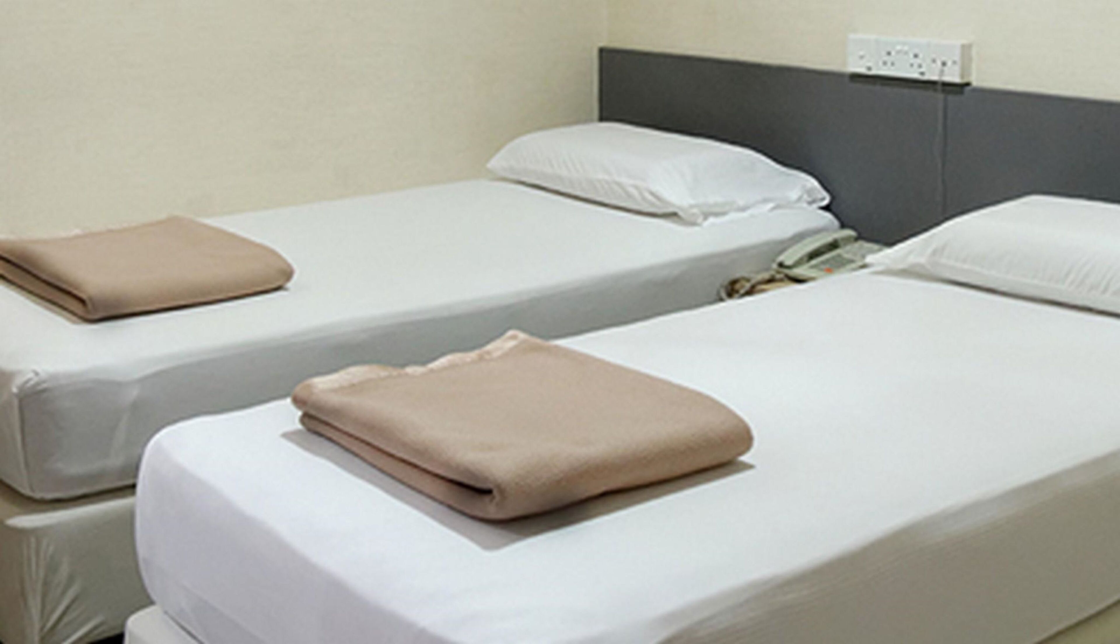 Twee enkele bedden met witte lakens en beige dekens, een grijs hoofdbord en een wit stopcontact aan de muur.