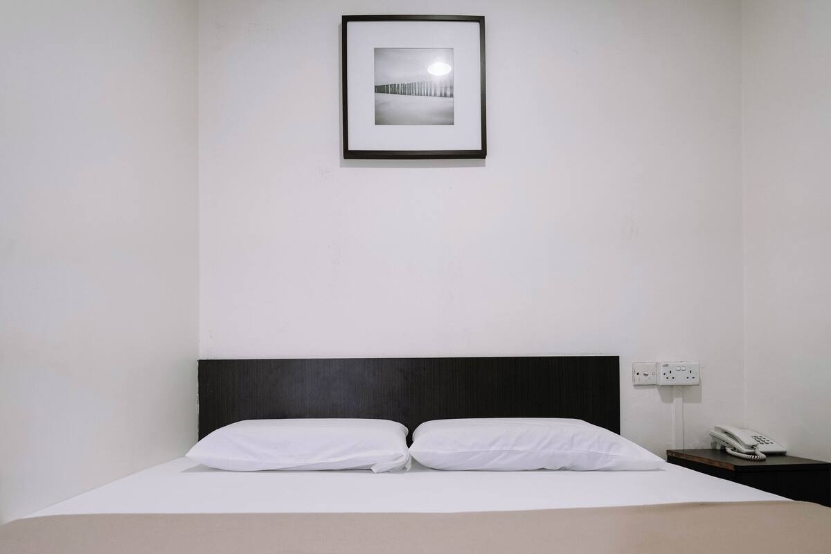 A neatly made bed with white linens, a framed picture above it, and a bedside phone on a wooden nightstand.