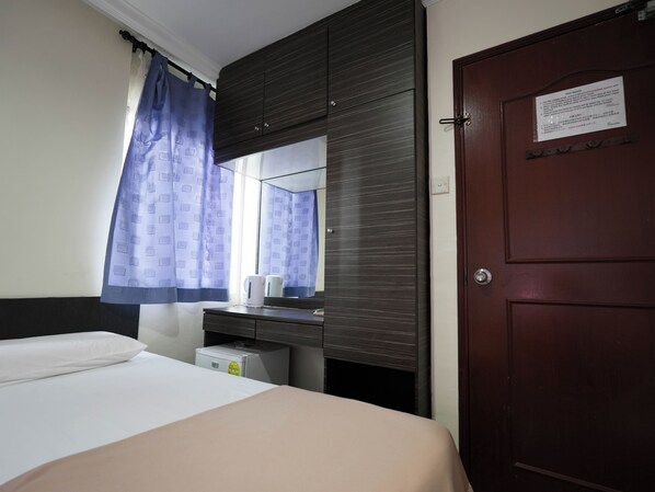 A hotel room with a bed, a small desk with a kettle, a mirror, and a door with a notice. - Amrise Hotel (Singapore)