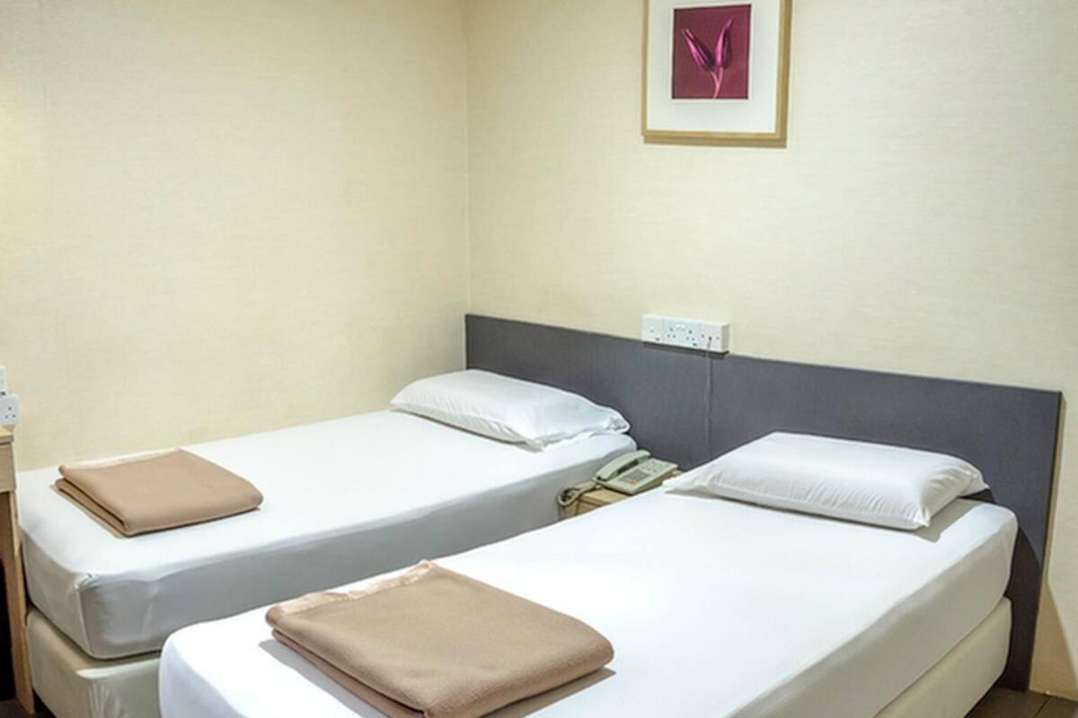 A hotel room with two single beds, each with a white mattress and a beige blanket. There is a framed picture on the wall above the beds.