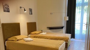 Ein Hotelzimmer mit zwei Betten, einem Heizkörper, einer Wandleuchte und einem mit Vorhängen versehenen Fenster.