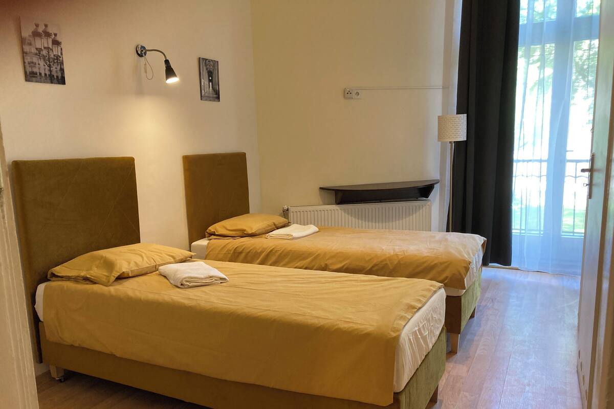 A hotel room with two beds, a radiator, a wall-mounted lamp, and a window with curtains.