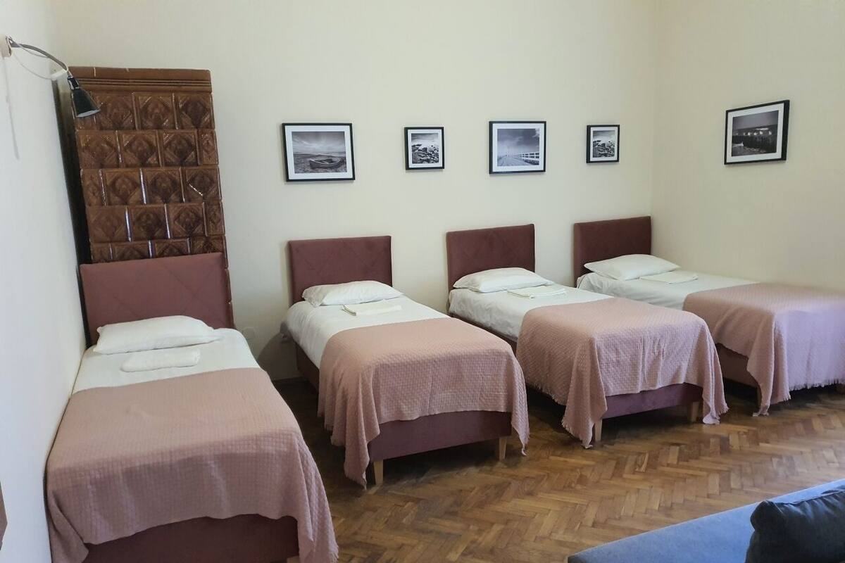 A room with four single beds, each with a pink bedspread and white pillows. There are four framed pictures on the wall, and a wooden herringbone floor.