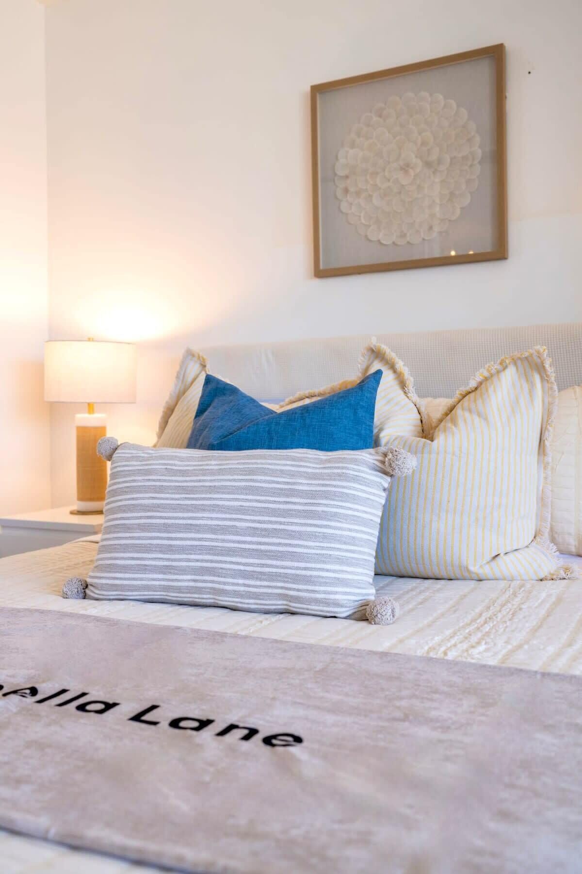 A neatly made bed with a blanket labeled "Allia Lane," a bedside lamp, and a framed artwork on the wall.