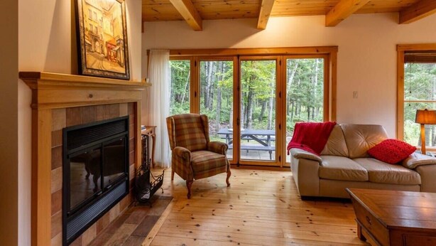 Una sala de estar acogedora con chimenea, un sillón a cuadros, un sofá con una manta roja y ventanas amplias que dan a un bosque.
