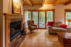 En hyggelig stue med pejs, en ternet lænestol, en sofa med rødt pled og store vinduer med udsigt til skoven.