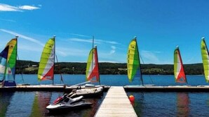 Un muelle con varios veleros y un jet ski, un cielo azul despejado y un lago tranquilo.