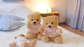 Deux ours en peluche sont posés sur un lit, accompagnés d’un oreiller à carreaux et d’une table de chevet sur laquelle se trouve une bougie.