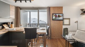 A modern kitchen with a dining area, a large window with a view of a snowy landscape, and a cozy living area with a sofa and a TV.