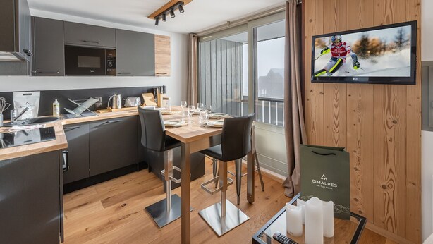 A modern kitchen with a dining area, a TV mounted on a wooden wall, and a view of the outdoors.