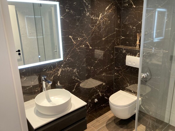 Une salle de bain moderne avec un pare-baignoire en marbre, un lavabo blanc, des toilettes et une douche à parois de verre.