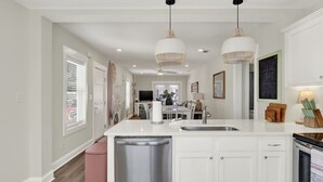 A modern kitchen with white cabinetry, a central island with a sink, pendant lighting, and a view into the dining area. - Brand New Tybee Beach Home: Walk to Beach (Unit A) (Tybee Island)
