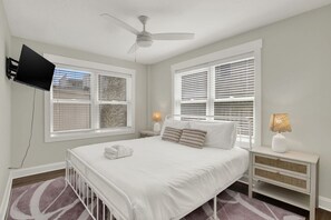 A bedroom with a bed, a ceiling fan, a TV mounted on the wall, and two windows with blinds.