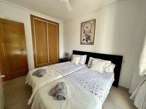 A double bed with a white and floral patterned bedspread, a headboard, and two pillows. There is a wooden door and a wall-mounted artwork. - Pool views apartment - AO112LT (La Torre Golf Resort)
