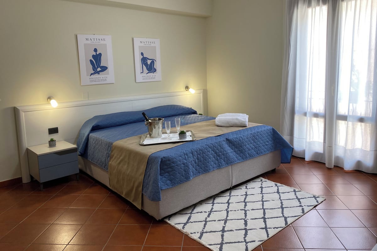 A neatly made bed with a blue quilt, a bedside table with a lamp, and two framed artworks on the wall.