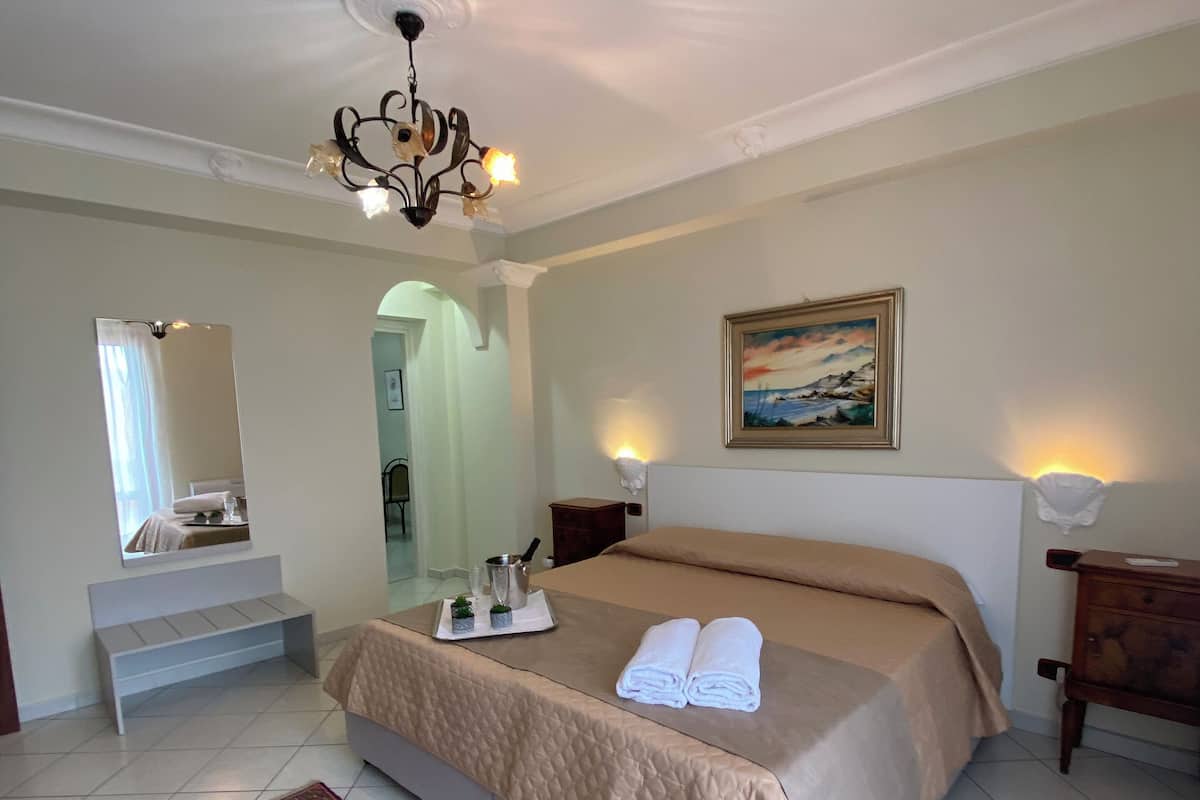 A hotel room with a bed, a large mirror, a chandelier, a painting, and a patterned rug.