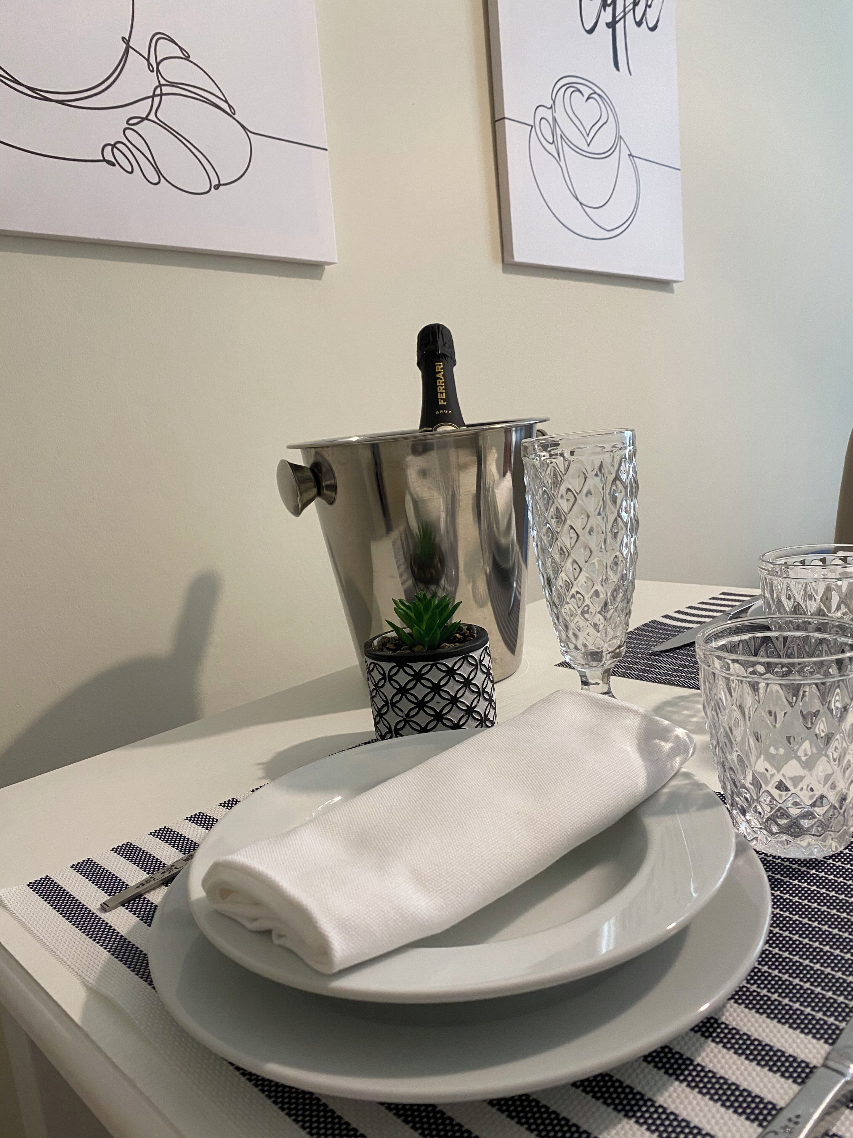 Une table avec une assiette blanche, une serviette blanche pliée, une petite plante en pot, un pichet métallique et deux verres transparents.