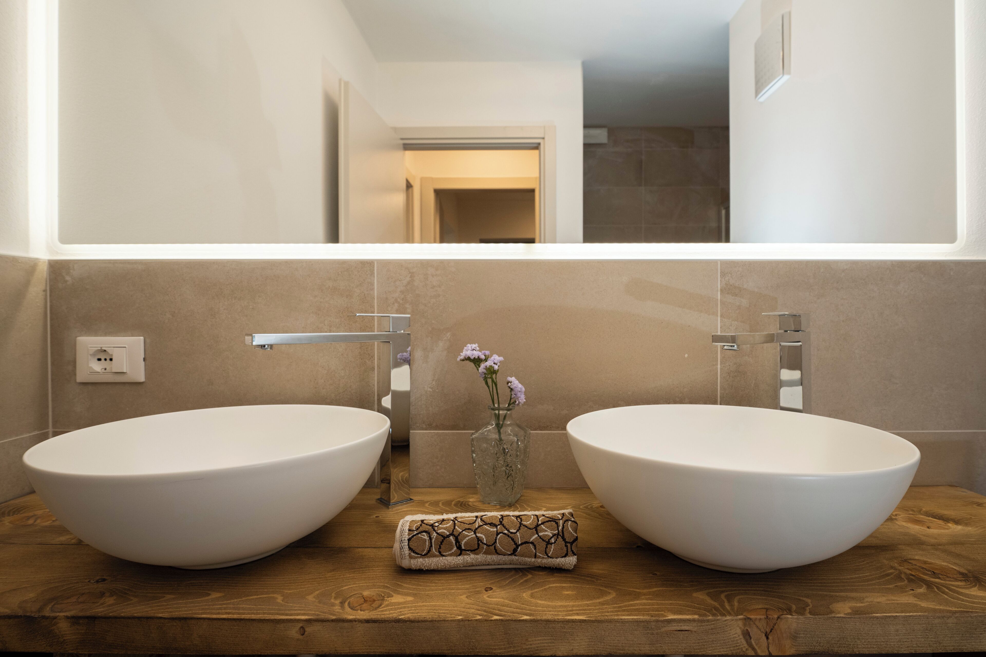 A bathroom with two large white vessel sinks, a wooden countertop, a small vase with flowers, and a mirror above the sinks.