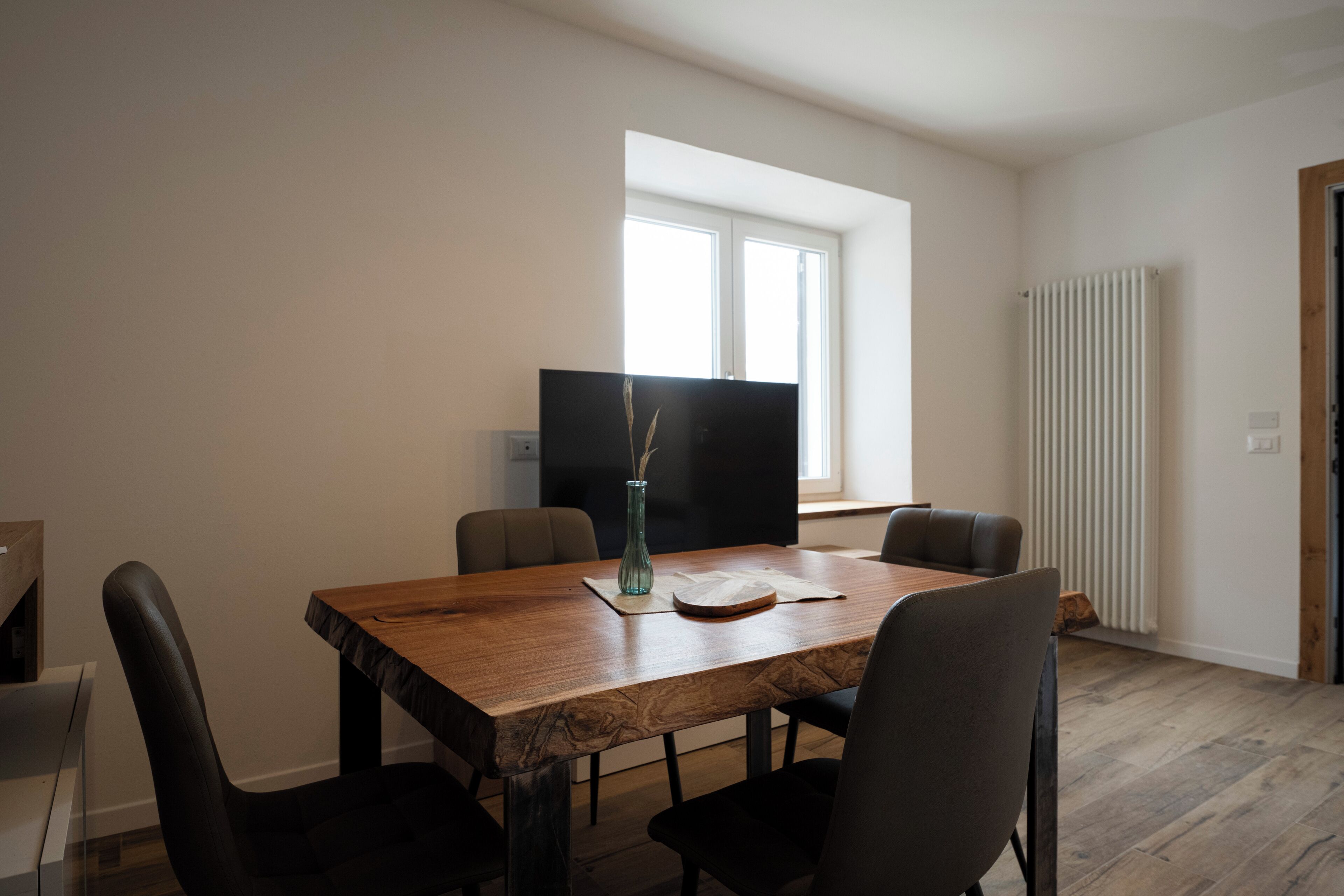 Un comedor con mesa de madera, cuatro sillas, un florero y un televisor de pantalla plana.