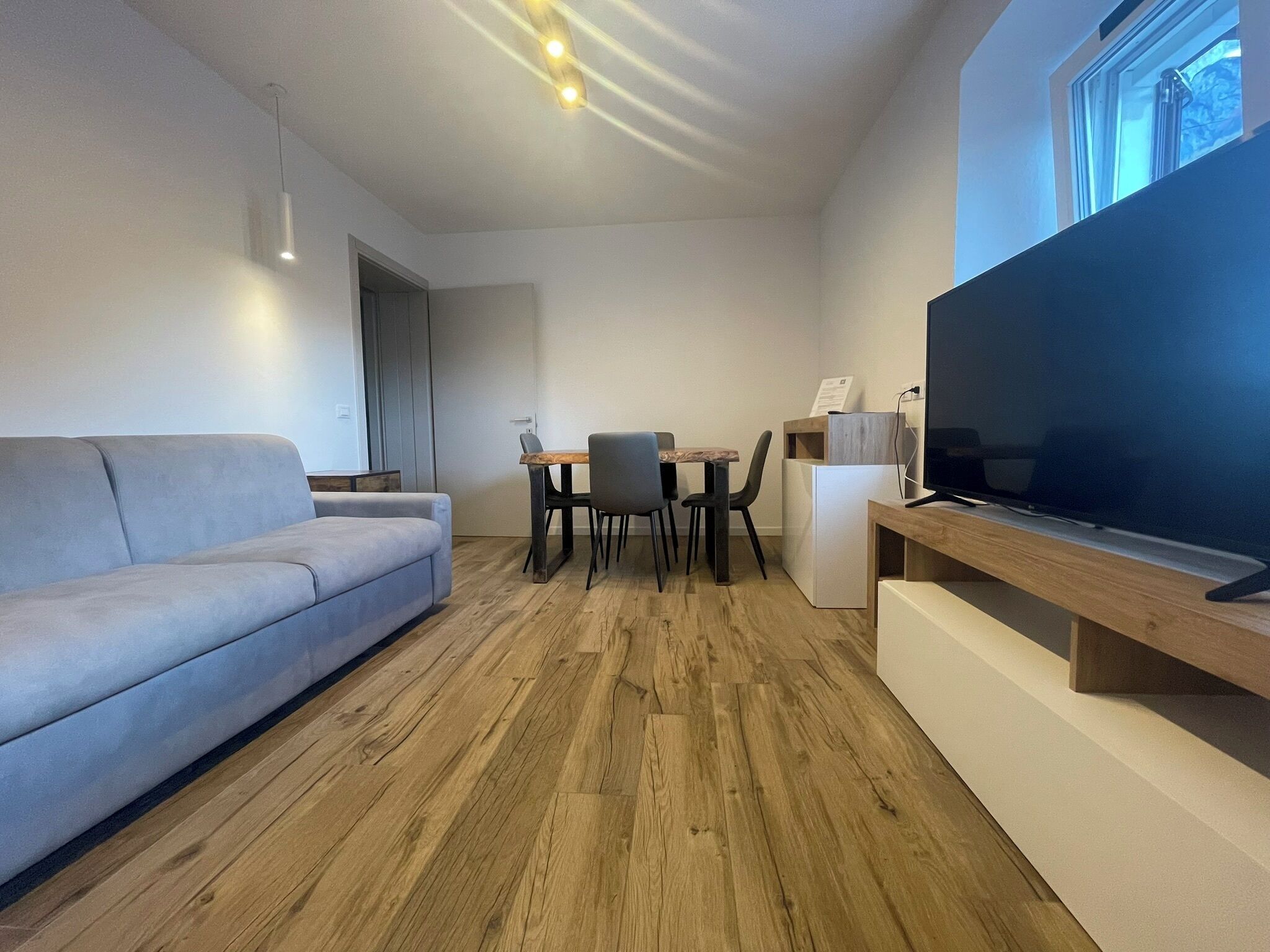 Una sala de estar moderna con un sofá gris, piso de madera, un comedor con mesa y sillas, y un televisor de pantalla plana grande.