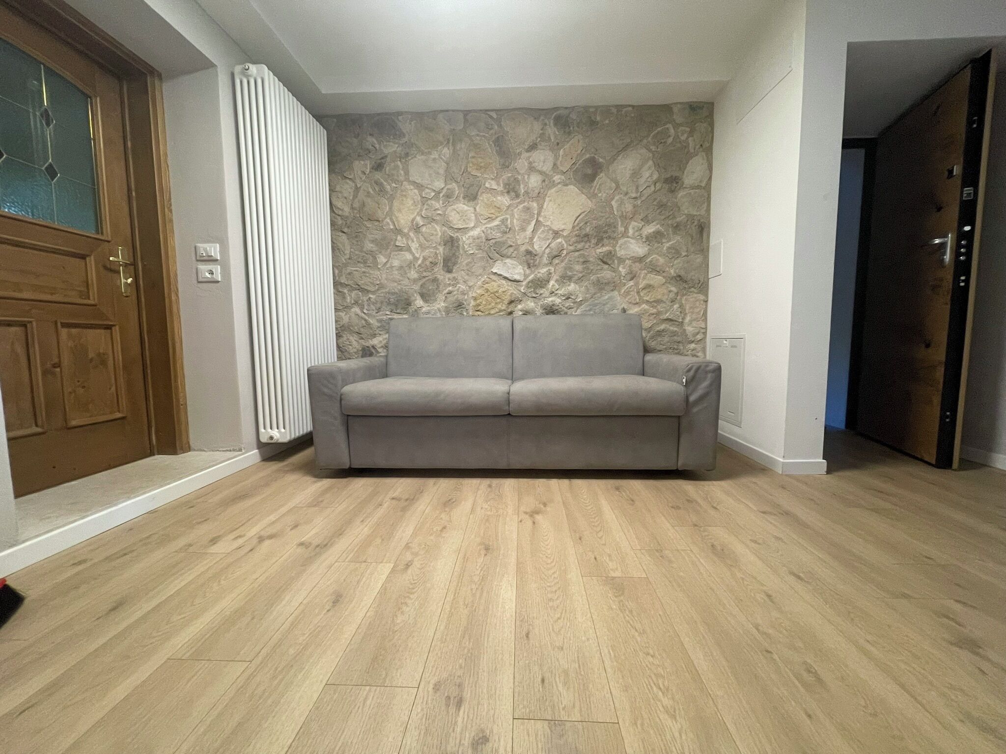 Una habitación con pared de piedra, un sofá gris y suelo de madera.