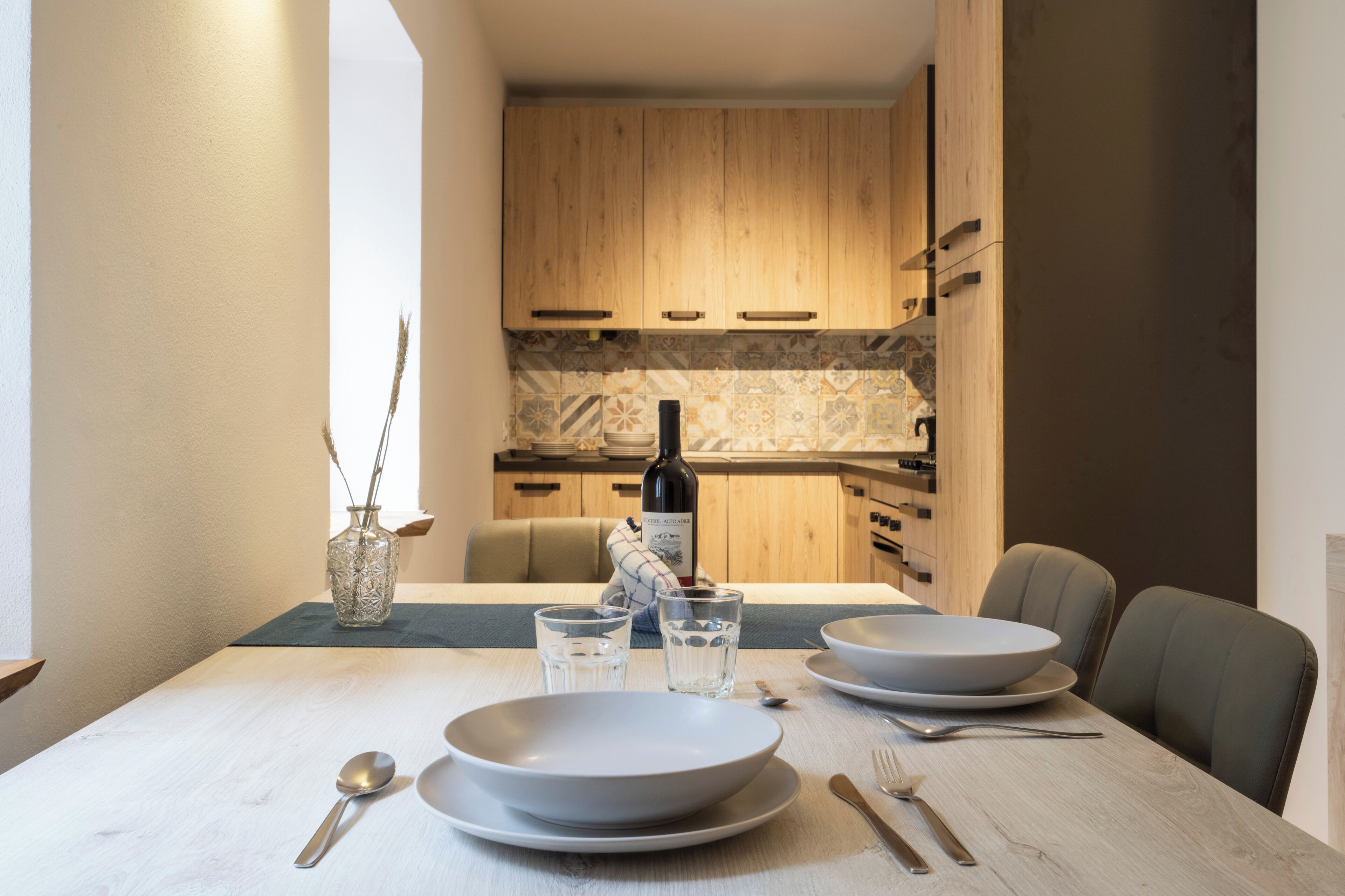 Een eettafel gedekt met borden, kommen, glazen en bestek in een moderne keuken met houten kastjes en een betegelde achterwand.