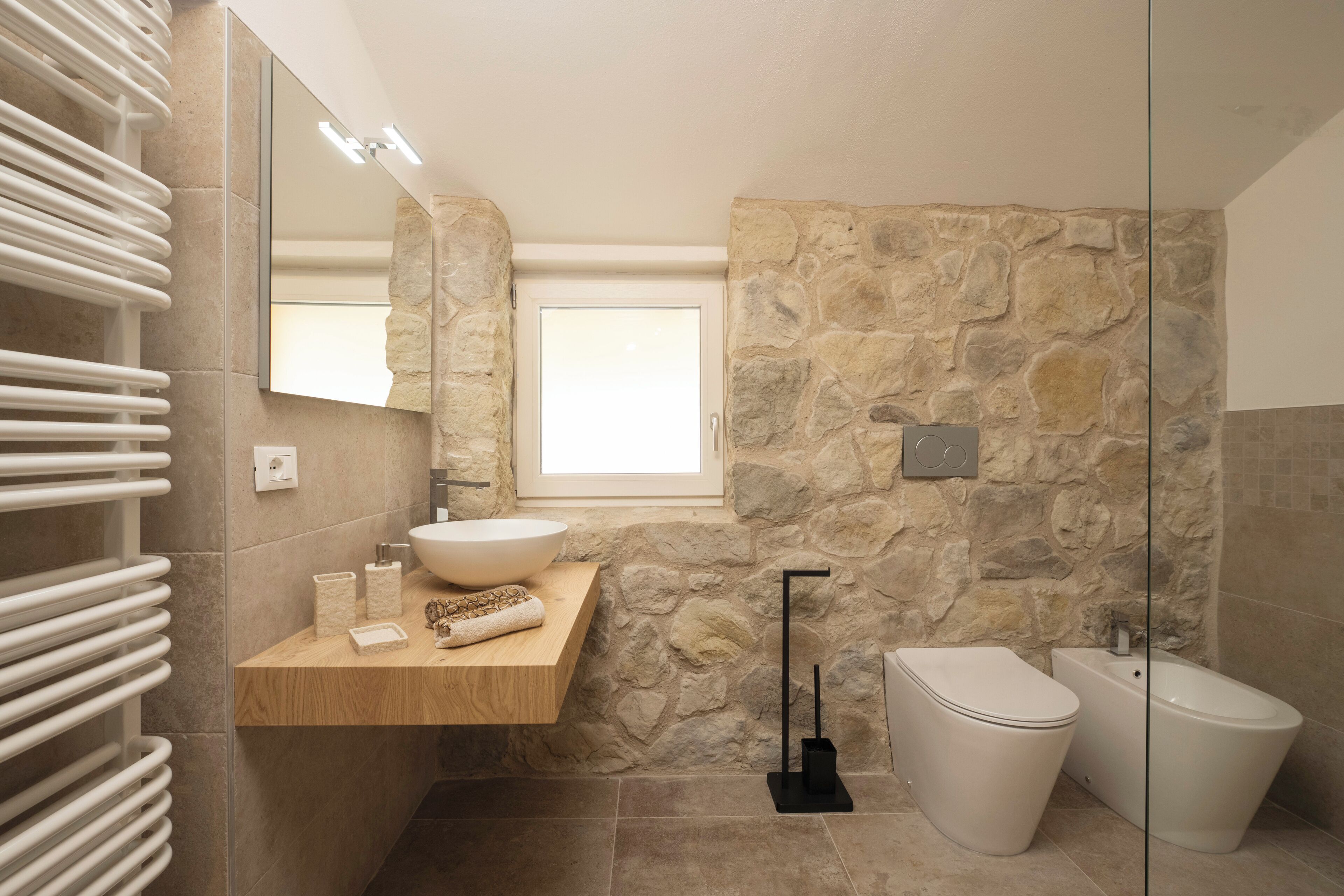 Ein modernes Badezimmer mit Steinwand, Waschtisch aus Holz, großem Spiegel, weißem Waschbecken, Toilette und Bidet.