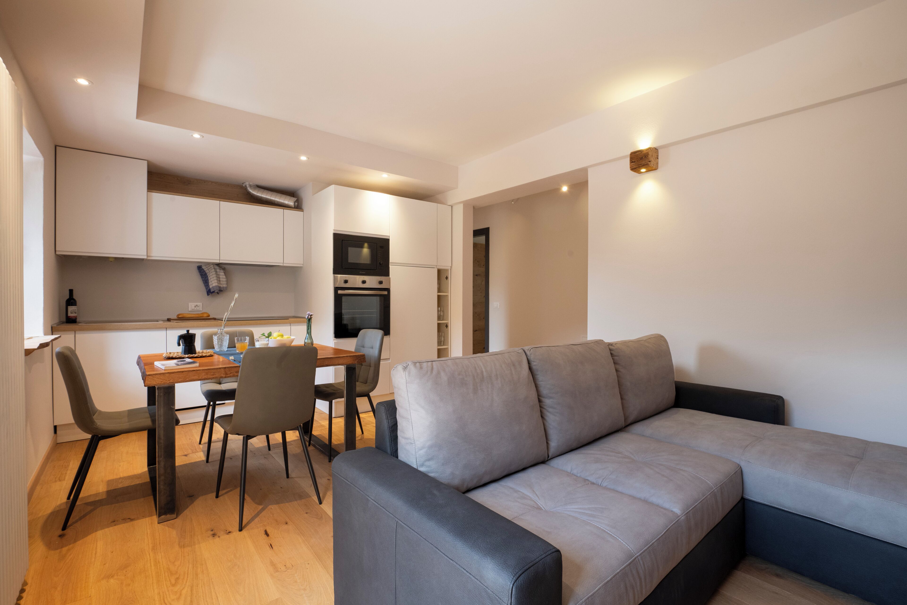 A modern living space with a grey sofa, wooden dining table, and chairs. There is a kitchen area with cabinets and appliances.