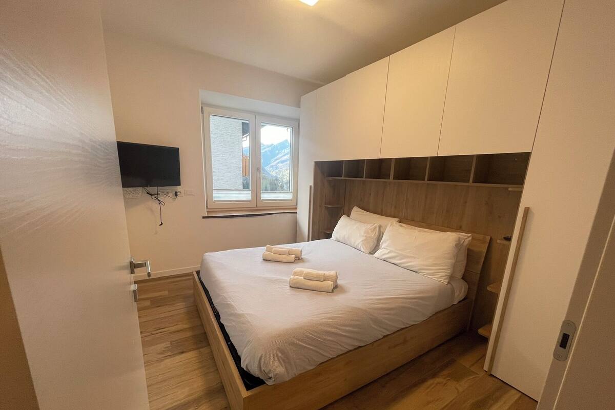 Ein Schlafzimmer mit einem hölzernen Bett, weißer Bettwäsche, einem Fenster mit Blick auf die Berge und einem an der Wand montierten Flachbildfernseher.