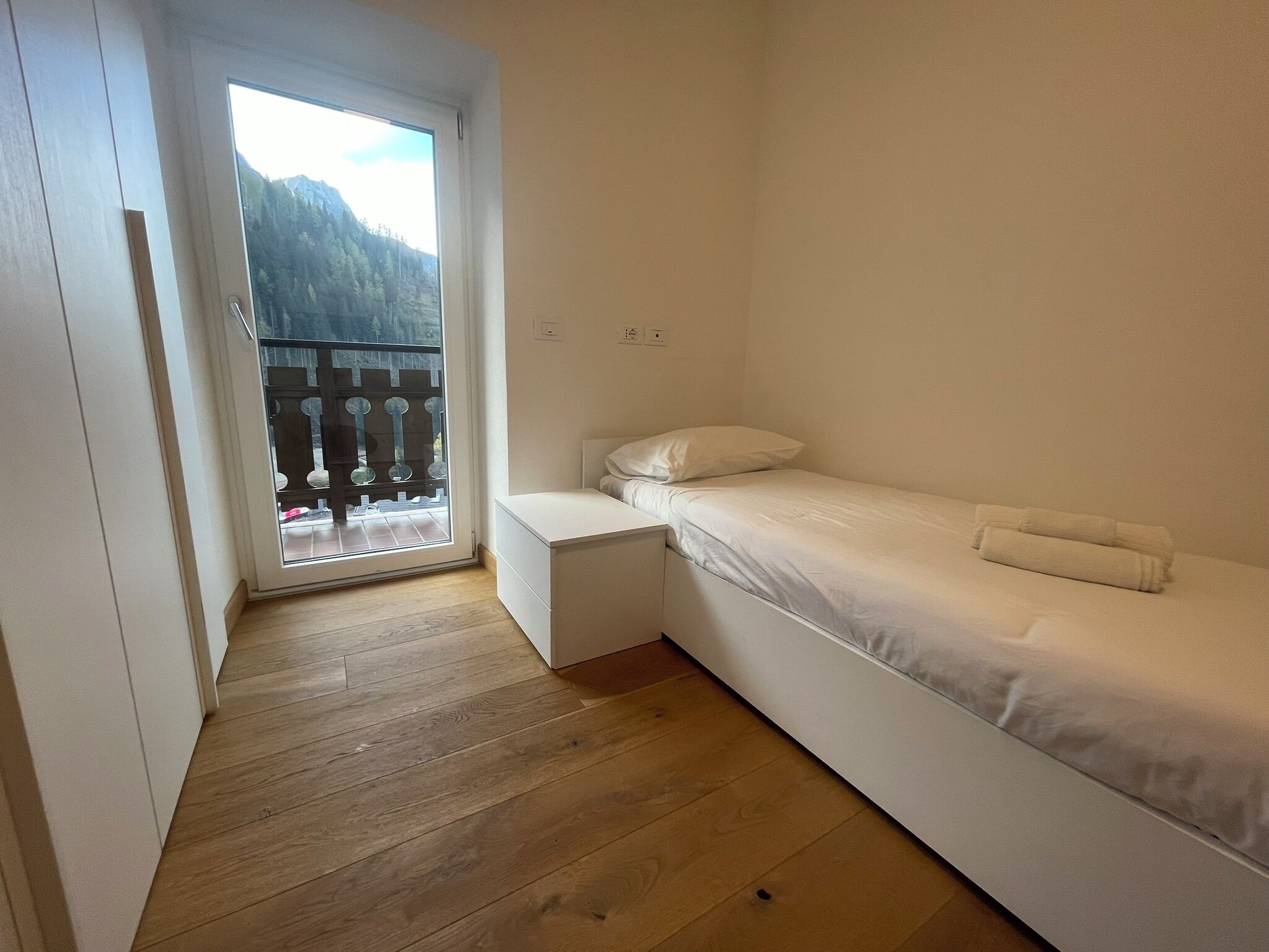 A small, single-bed room with a wooden floor, a white bed, and a balcony with a view of a mountain.