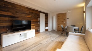A modern living room with a flat-screen TV on a white entertainment unit, wooden flooring, a beige sofa, and a dining area with a table and chairs. - SnowLake Suite & Apartments (Alleghe)