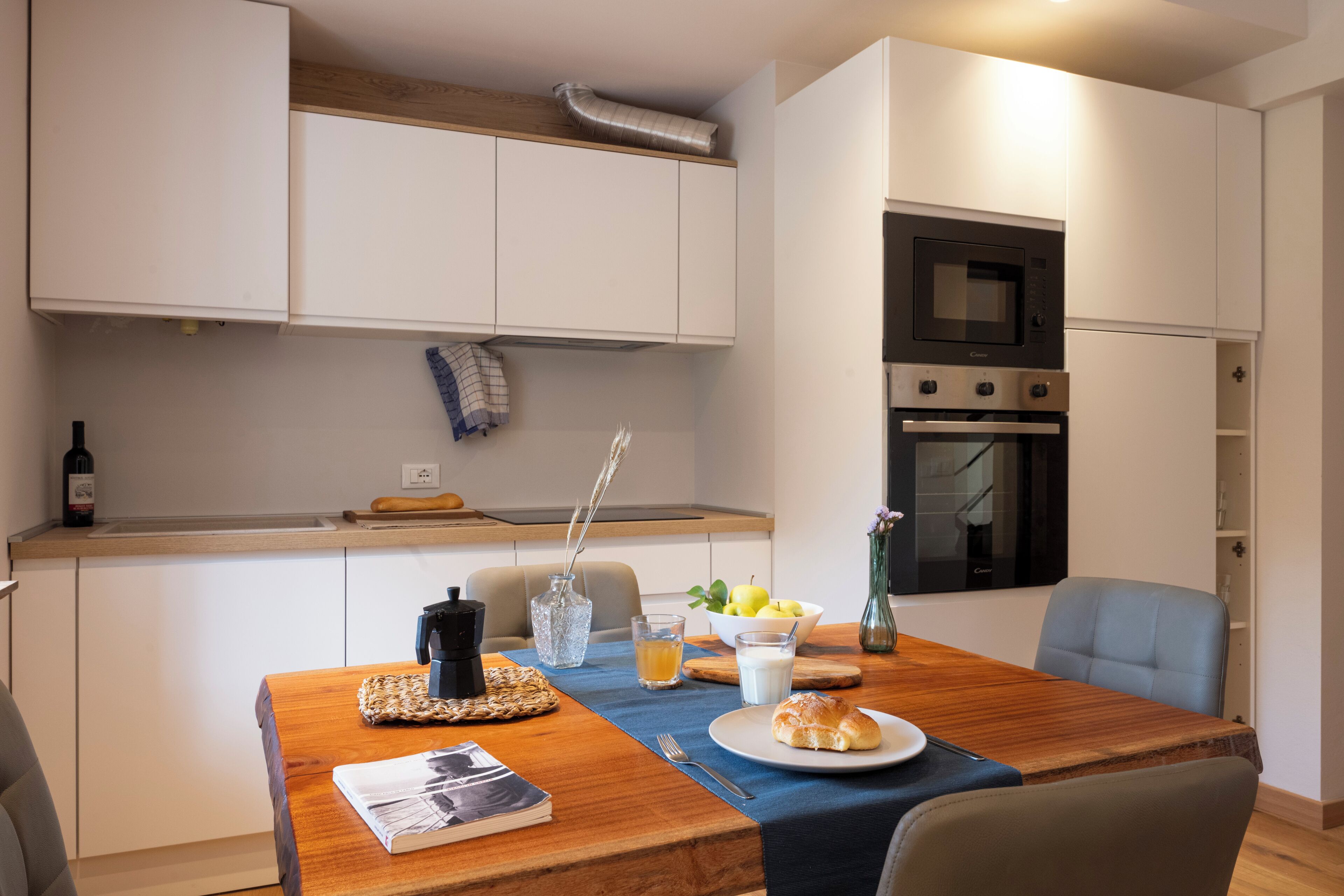 A modern kitchen with white cabinets, a microwave, and an oven. There is a dining table with a placemat, a plate with a croissant, and a bowl of fruit.