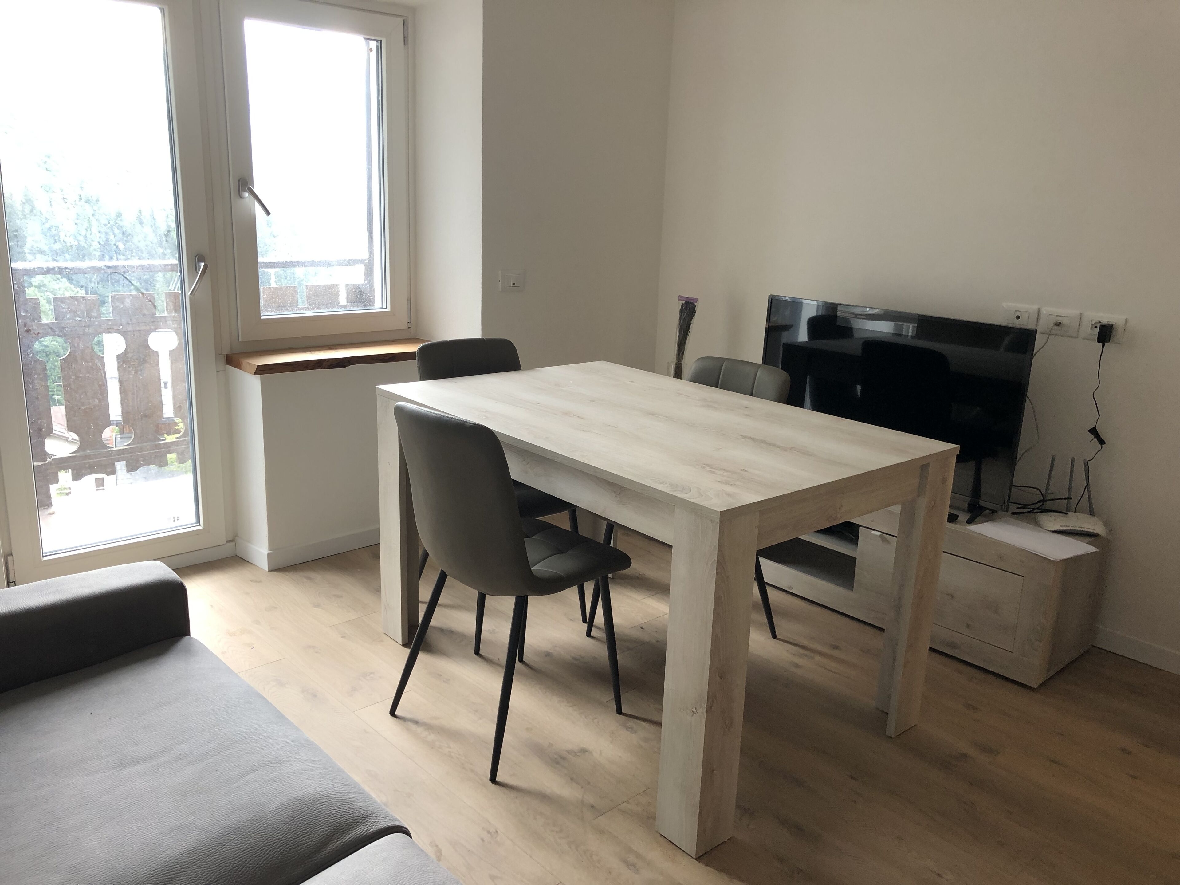 A modern, minimalist living space with a wooden dining table, two grey chairs, a grey sofa, and a TV on a stand.