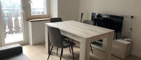 A modern, minimalist living space with a wooden dining table, two grey chairs, a grey sofa, and a TV on a stand.