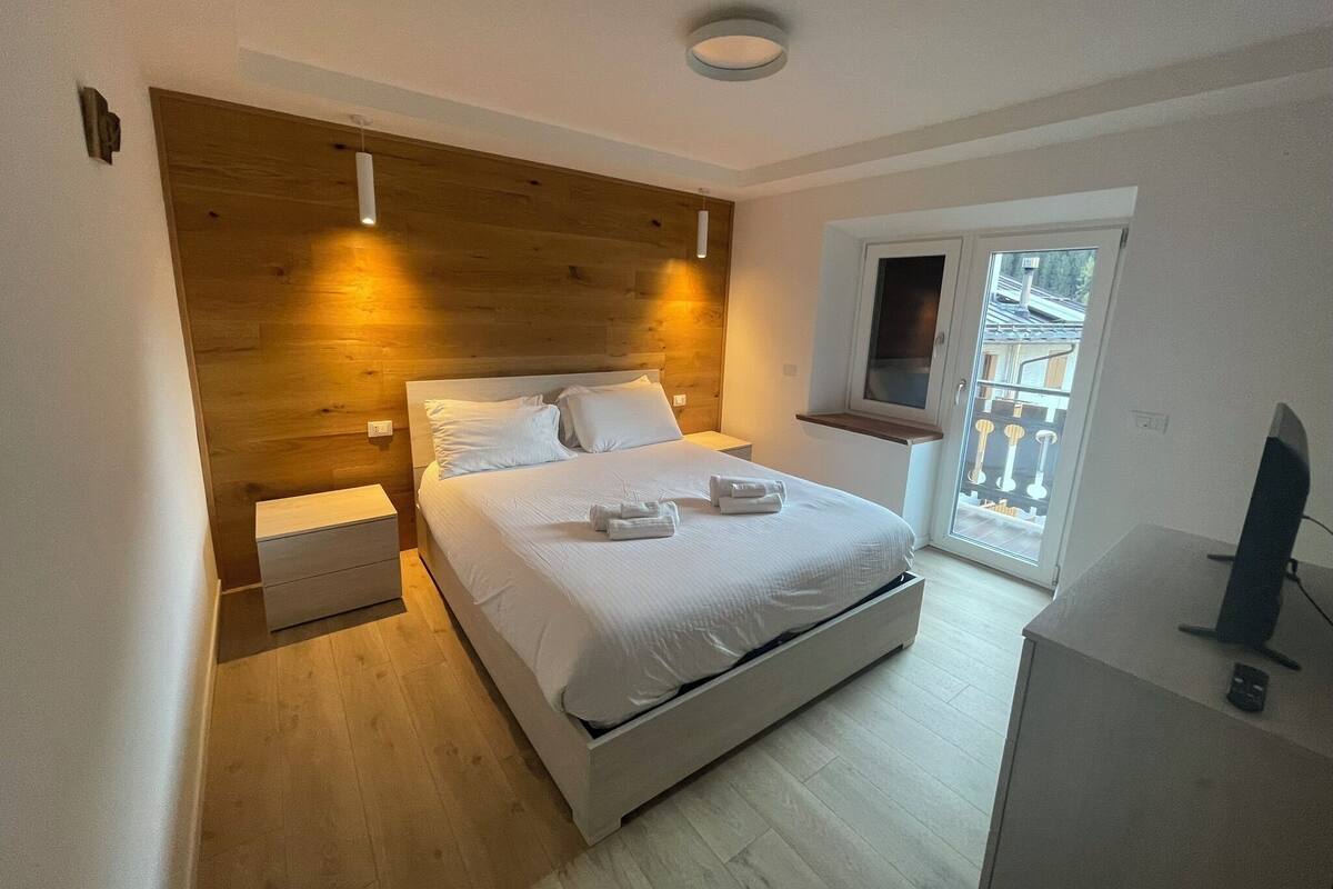 Ein Schlafzimmer mit massivem Kopfteil, einem Bett mit weißer Bettwäsche, einem Nachttisch und einem Fernseher auf einem Schrank.