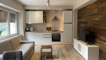 Een moderne keuken met witte kasten, een ingebouwde oven en een Nespresso machine. Er ligt een houten vloer en er staan een bank en een televisie op een wit meubel.