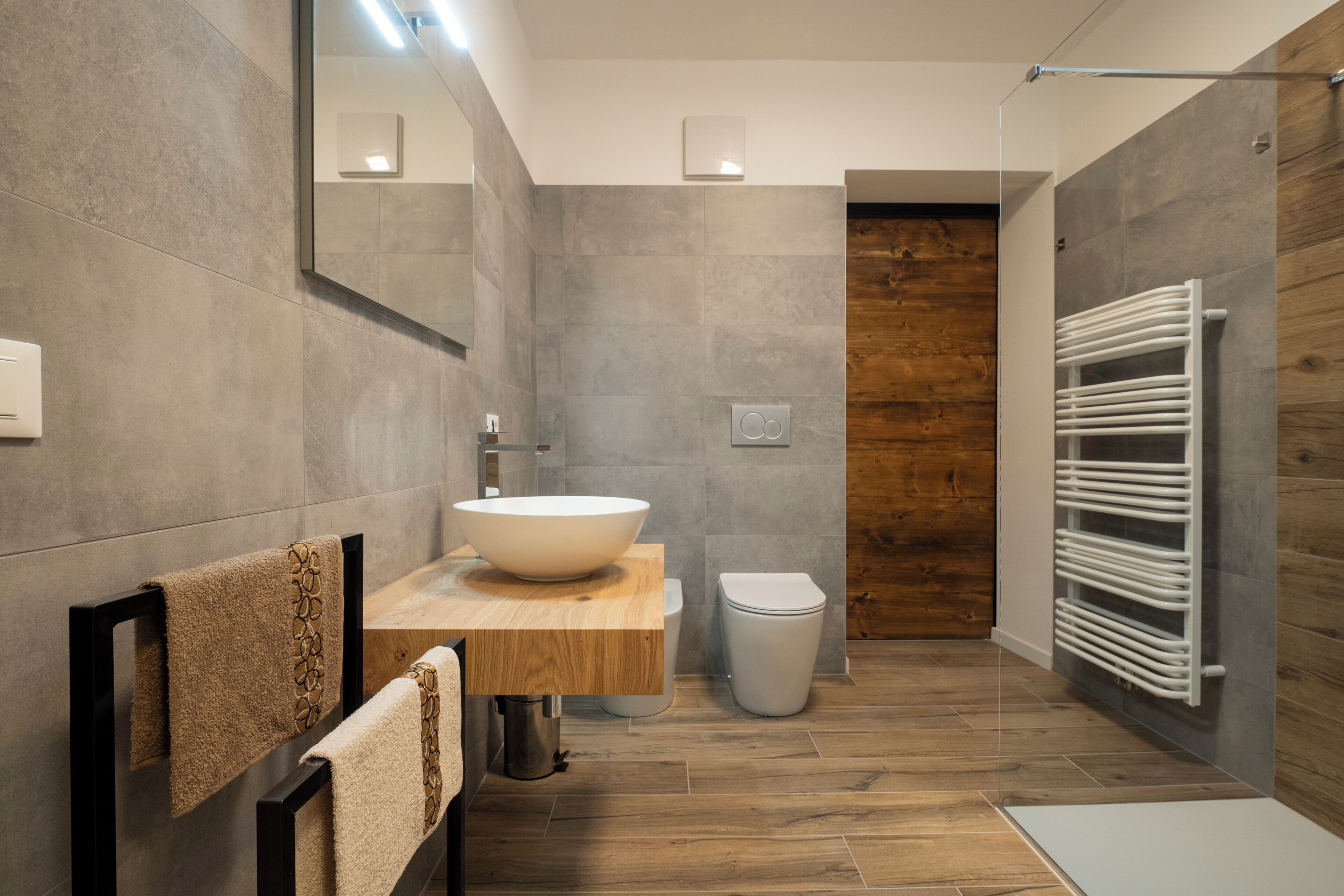 A modern bathroom with a large vessel sink, wooden vanity, a toilet, and a walk-in shower with a wooden wall.